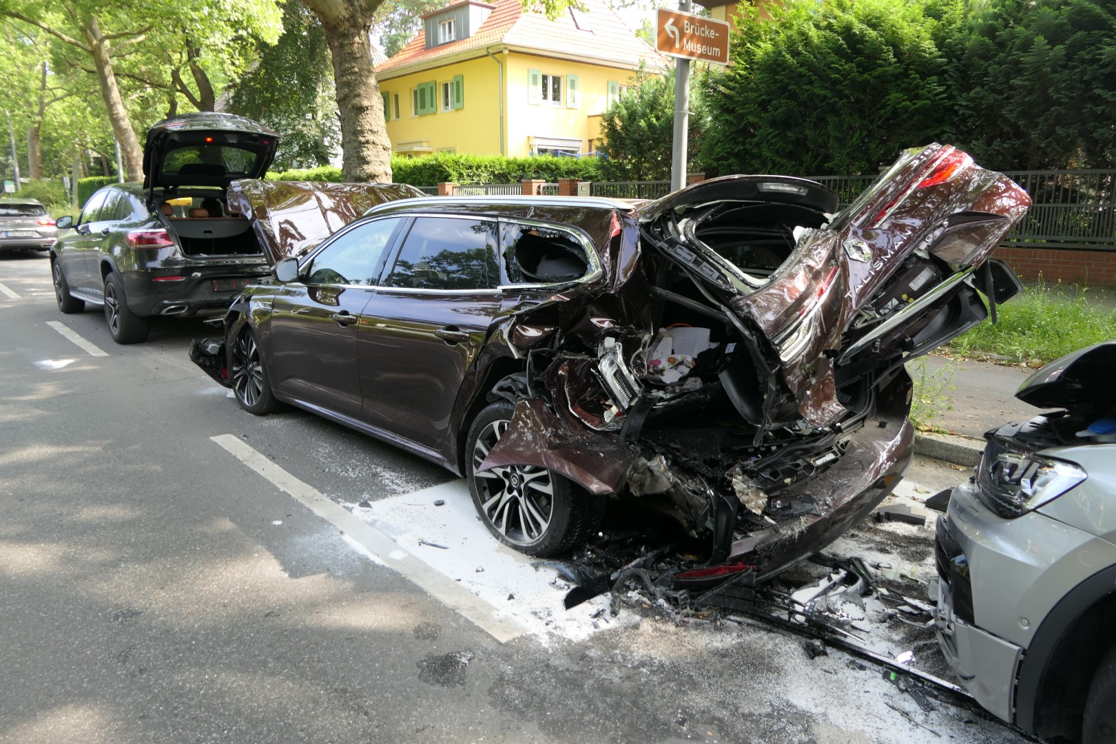 Verkehrsunfall mit drei beschädigten Fahrzeugen, parkend, sonniger Tag, Umgebung mit Bäumen.